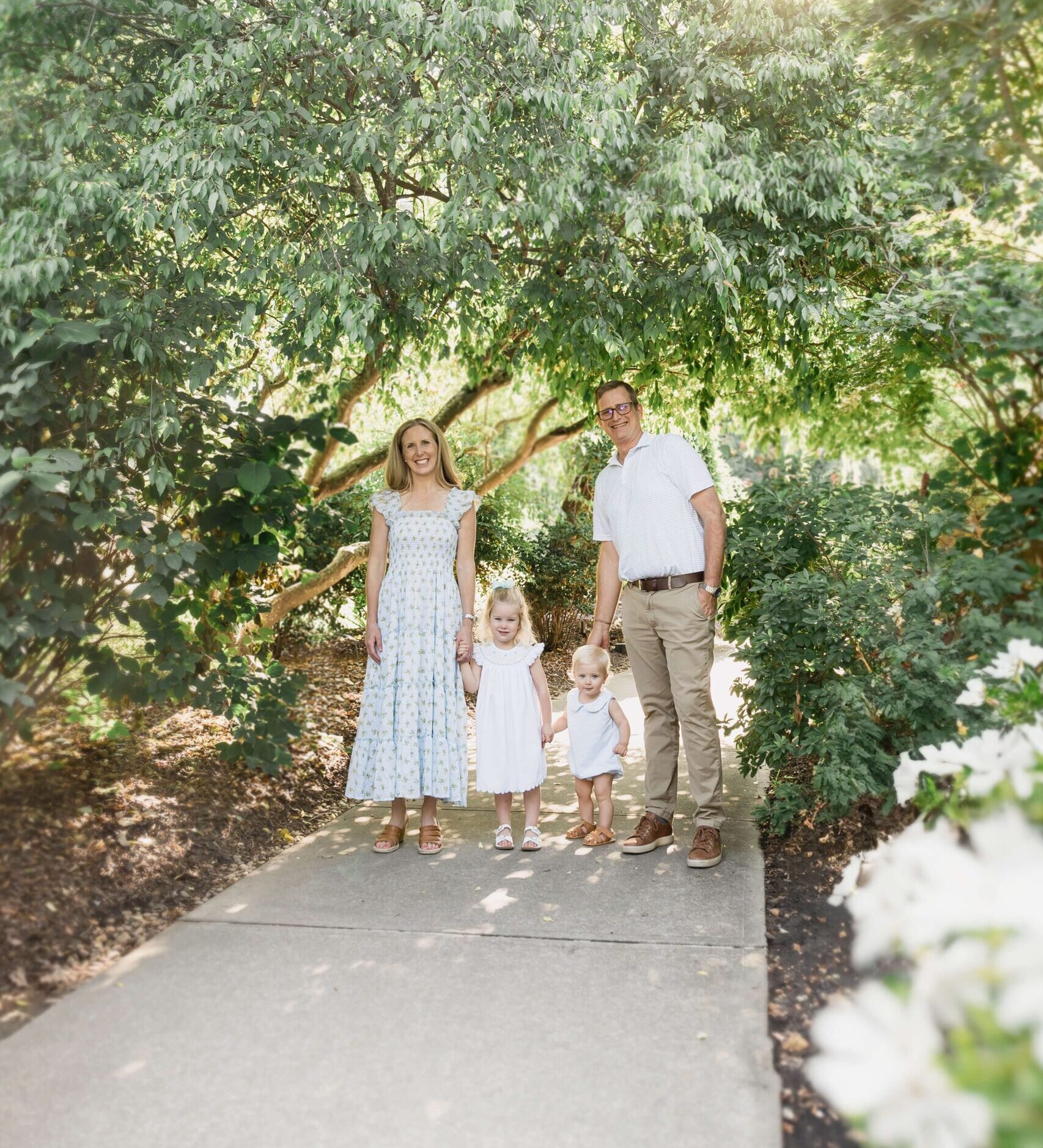 Toddler brother and sister hold hands with mom and dad while walking in a garden sidewalk