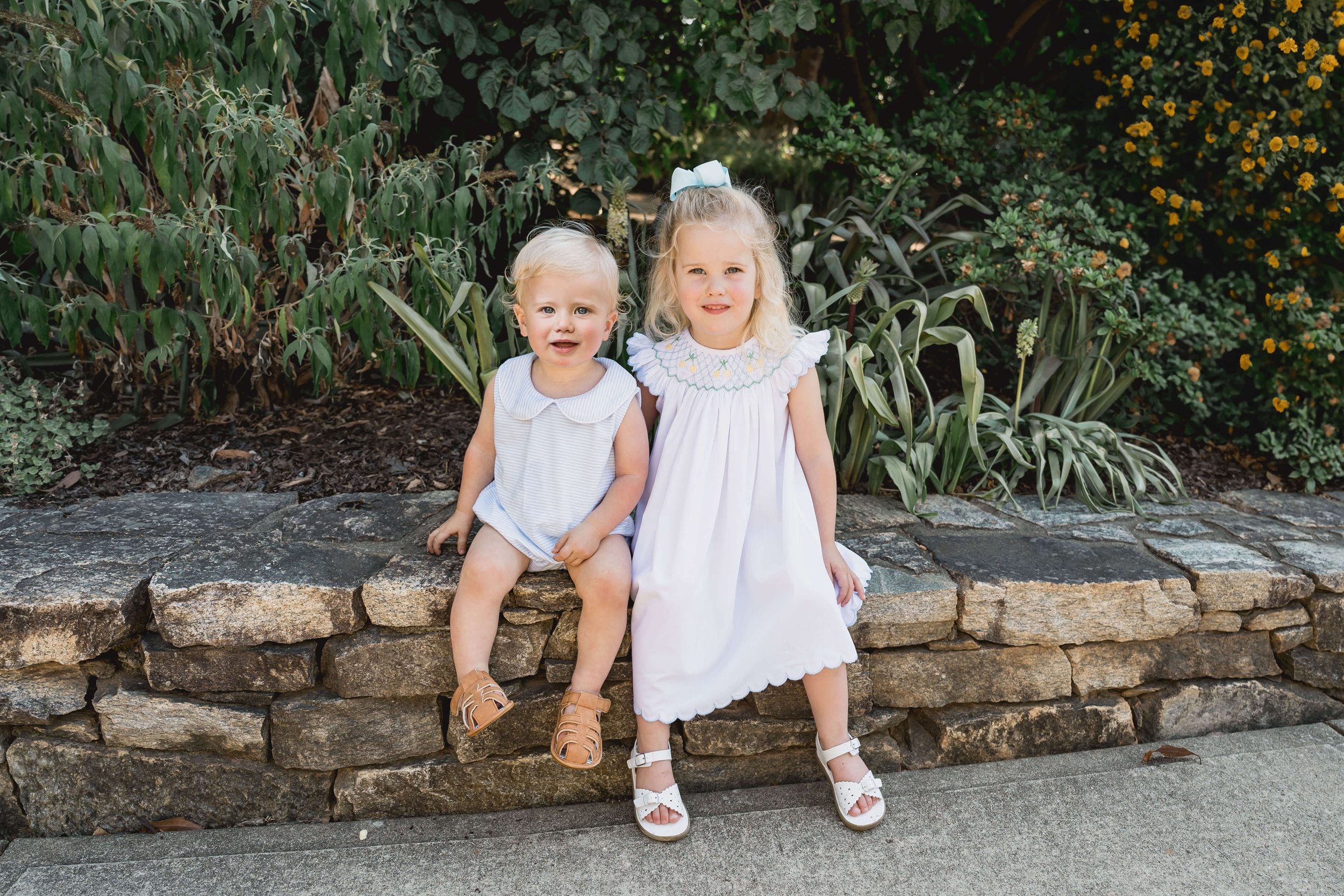 Happy toddler brother and sister sit on a stone garden wall in front of flowers