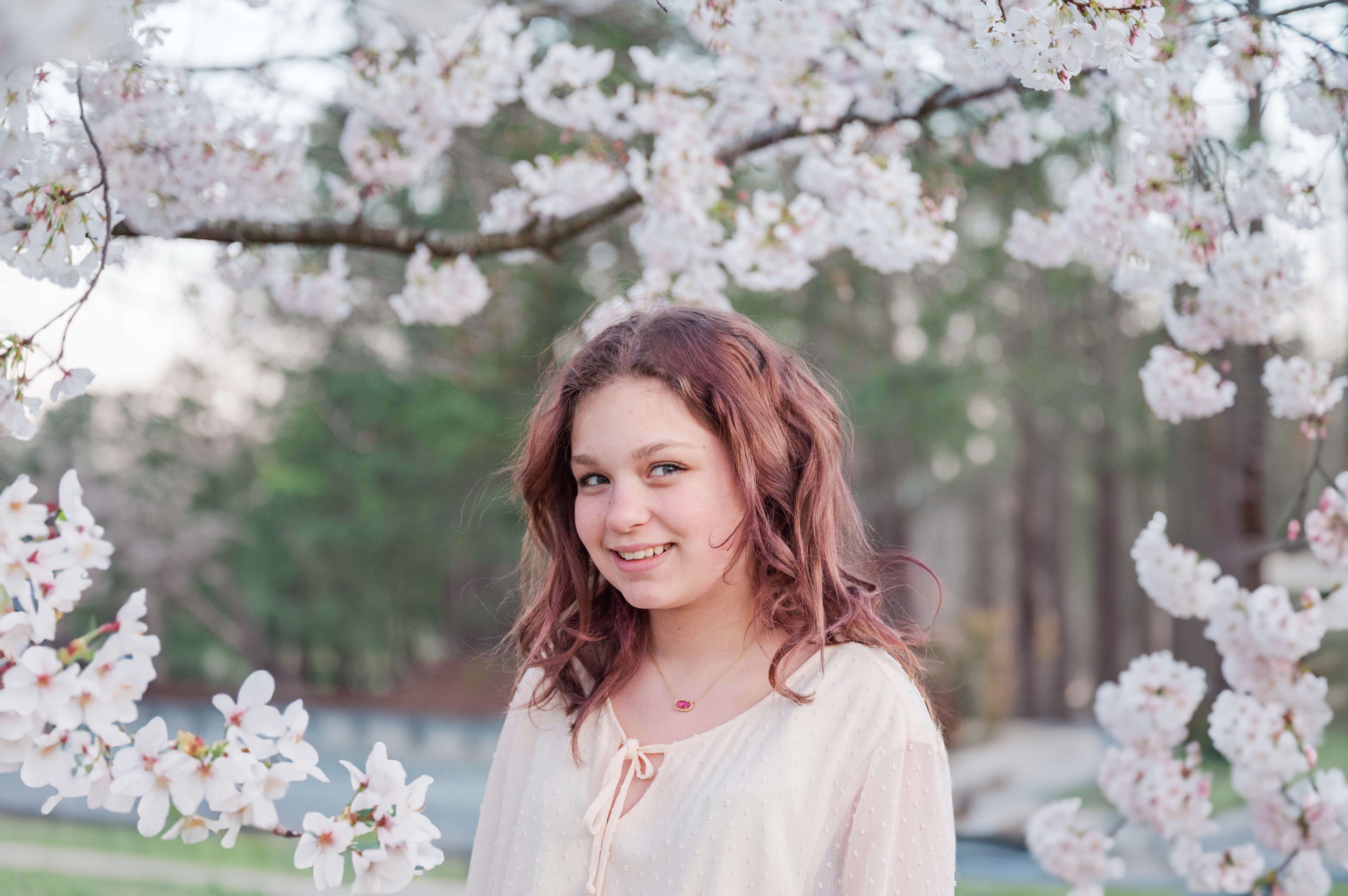 A young girl with red hair stands in a park under white blooming trees while smiling