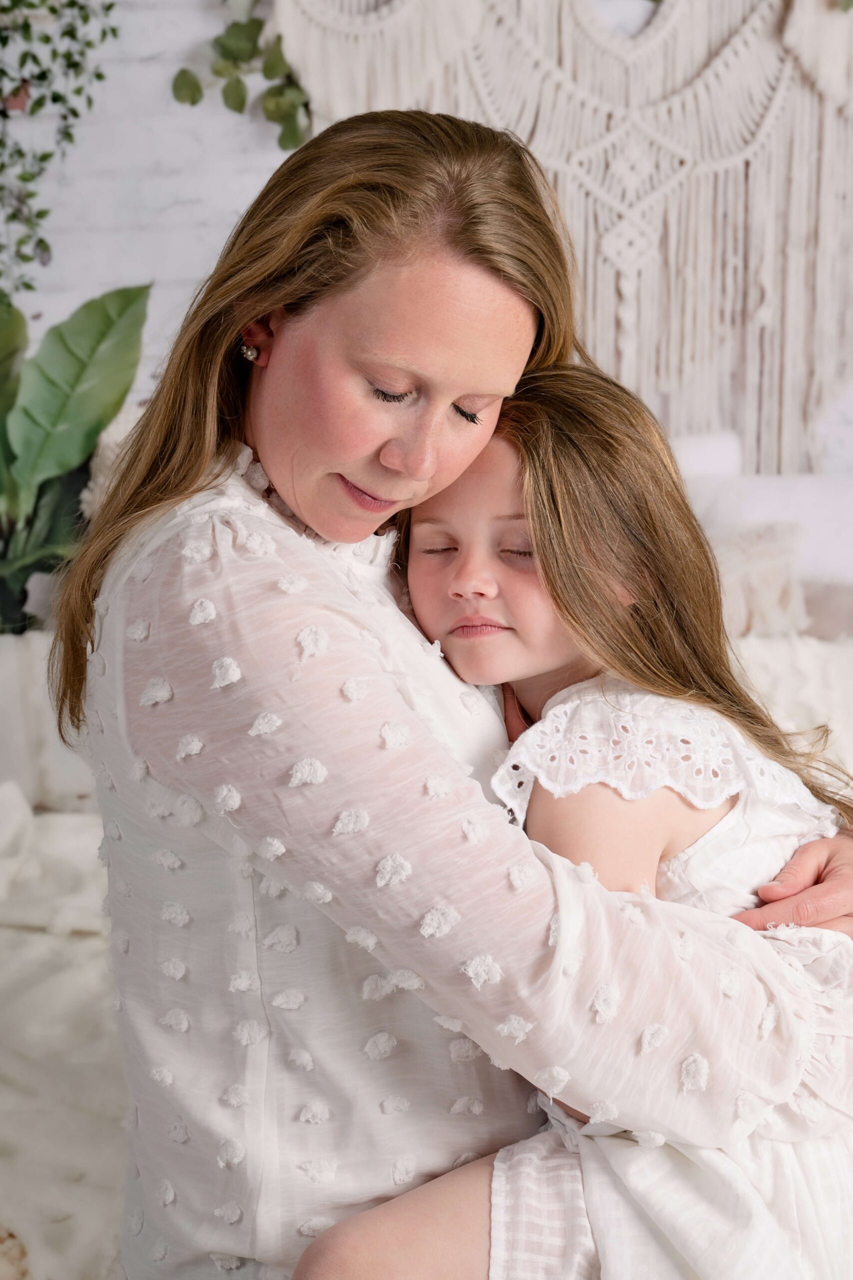 A mother and daughter sit on a bed hugging in white outfits after visiting harmony family chiropractic