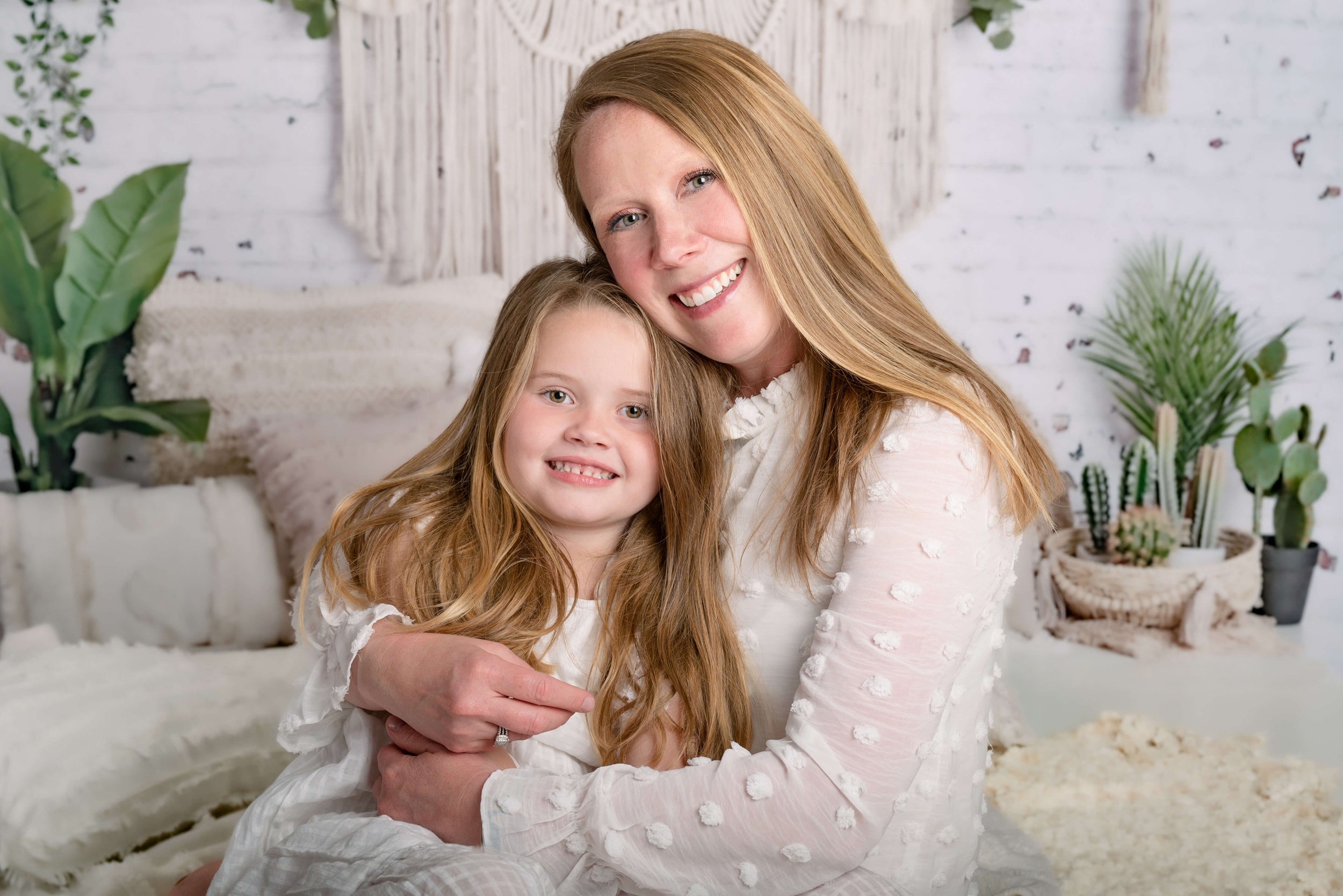 A happy mom sits on a bed hugging her toddler daughter in a white dress after visiting harmony family chiropractic