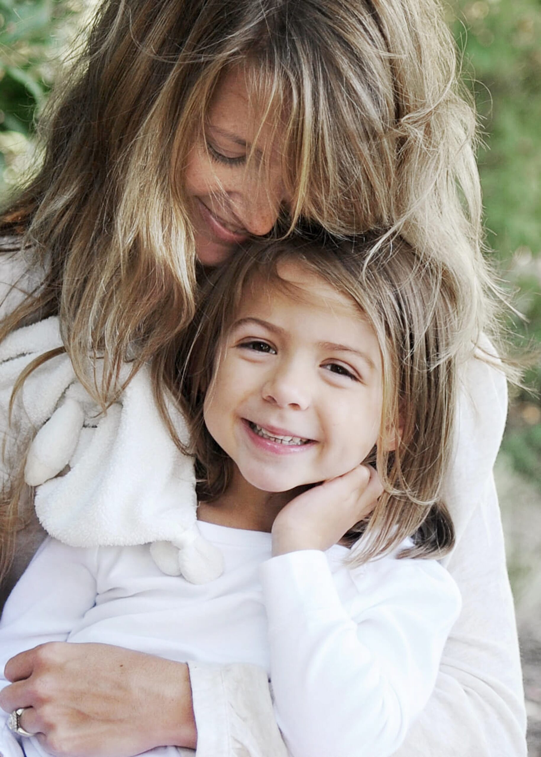 A happy toddler girl in a white shirt is hugged by mom in a garden