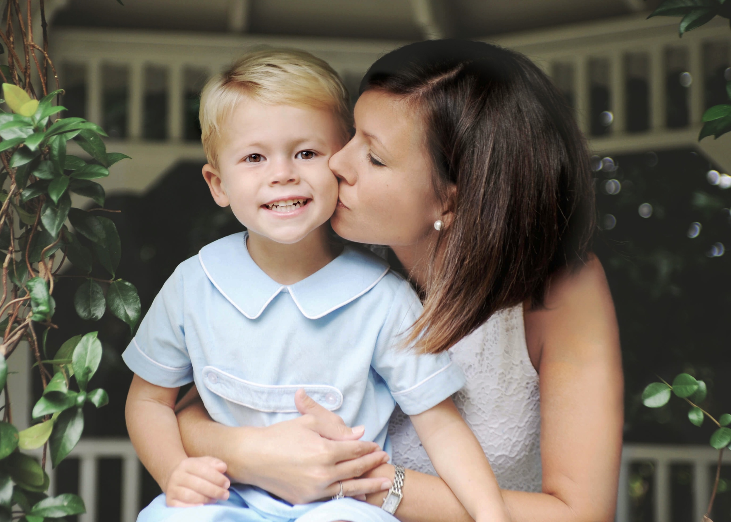 A happy toddler boy in a blue onesie sits on a gazebo railing while being hugged and kissed by mom before visiting pattywhacks