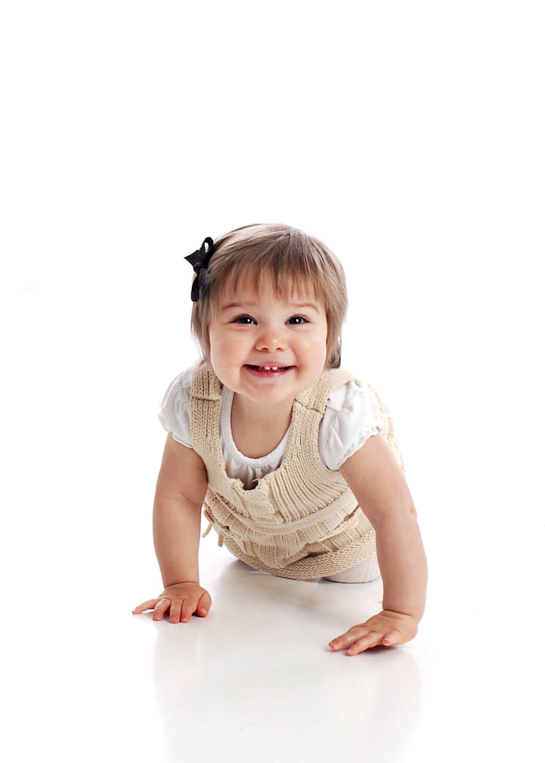 A smiling toddler girl in tan knit overalls crawls on the floor of a studio