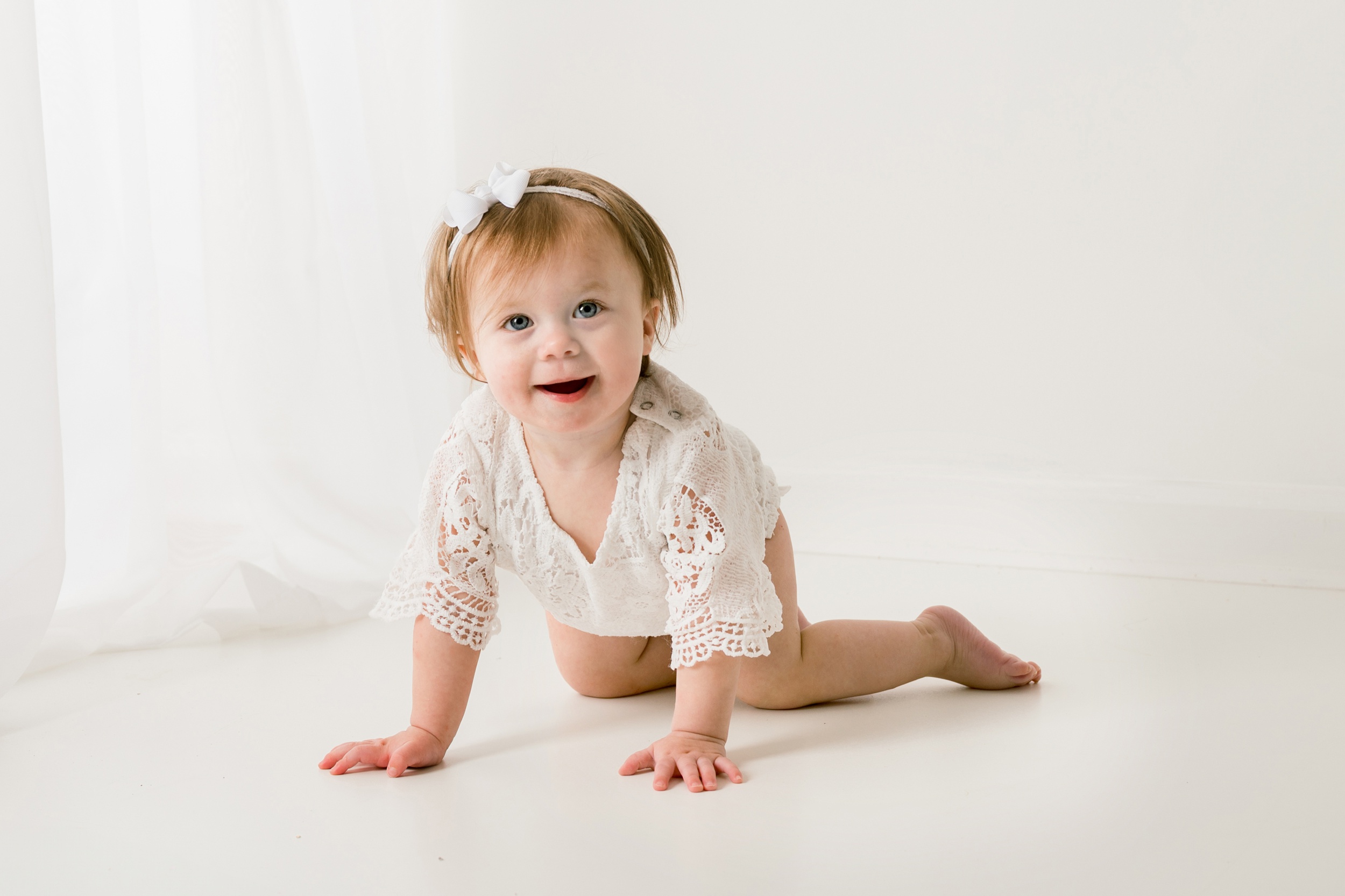 A toddler girl in a white lace onesie crawls on the floor of a studio before meeting pediatricians in raleigh nc