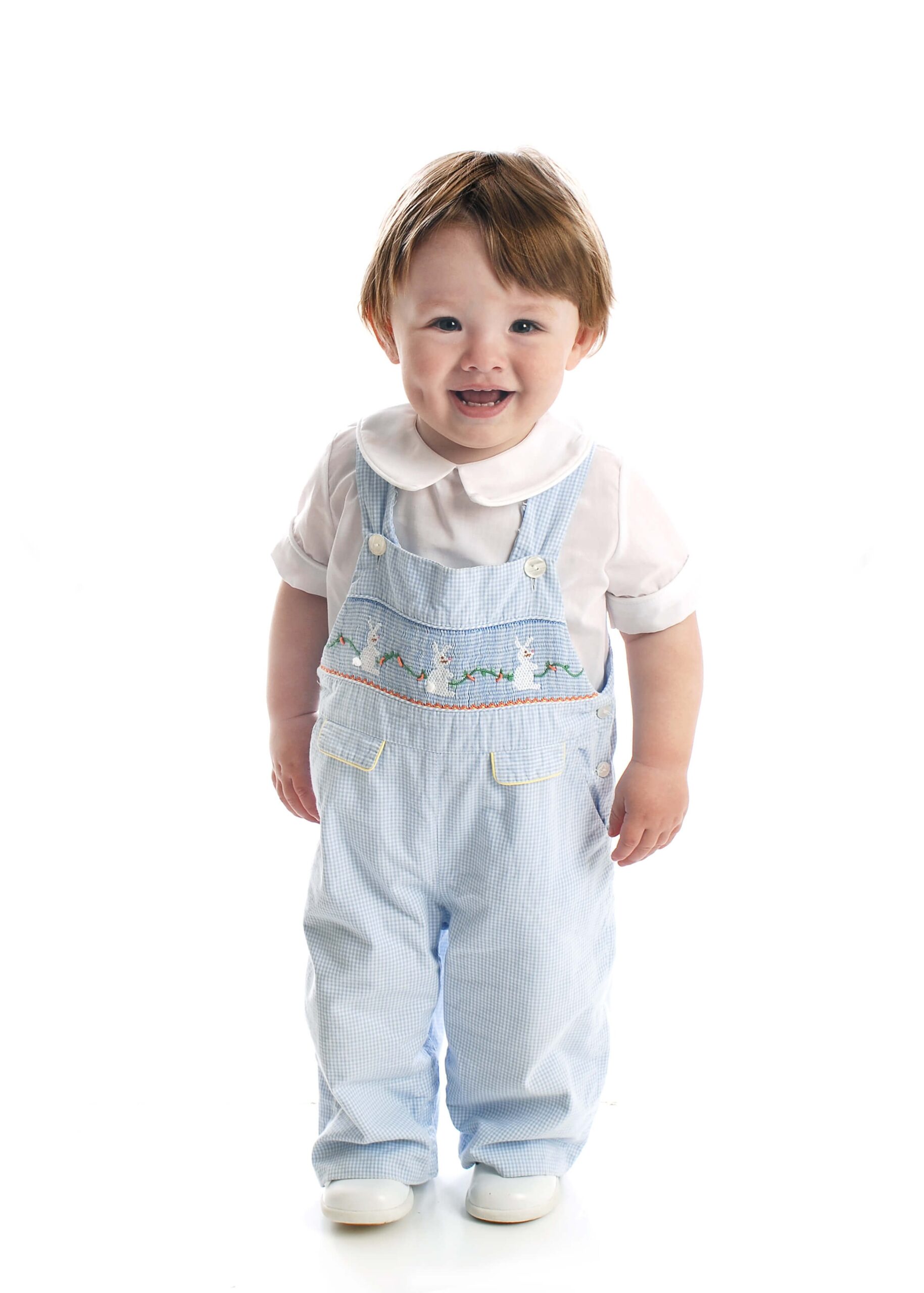 A happy toddler in blue easter bunny overalls stands in a studio smiling before visiting pediatricians in raleigh nc