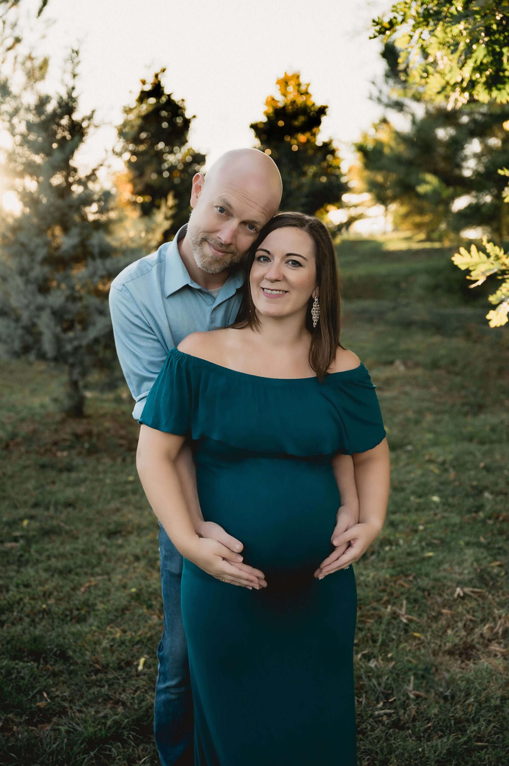 A happy mom to be is hugged from behind by her husband with hands on the bump