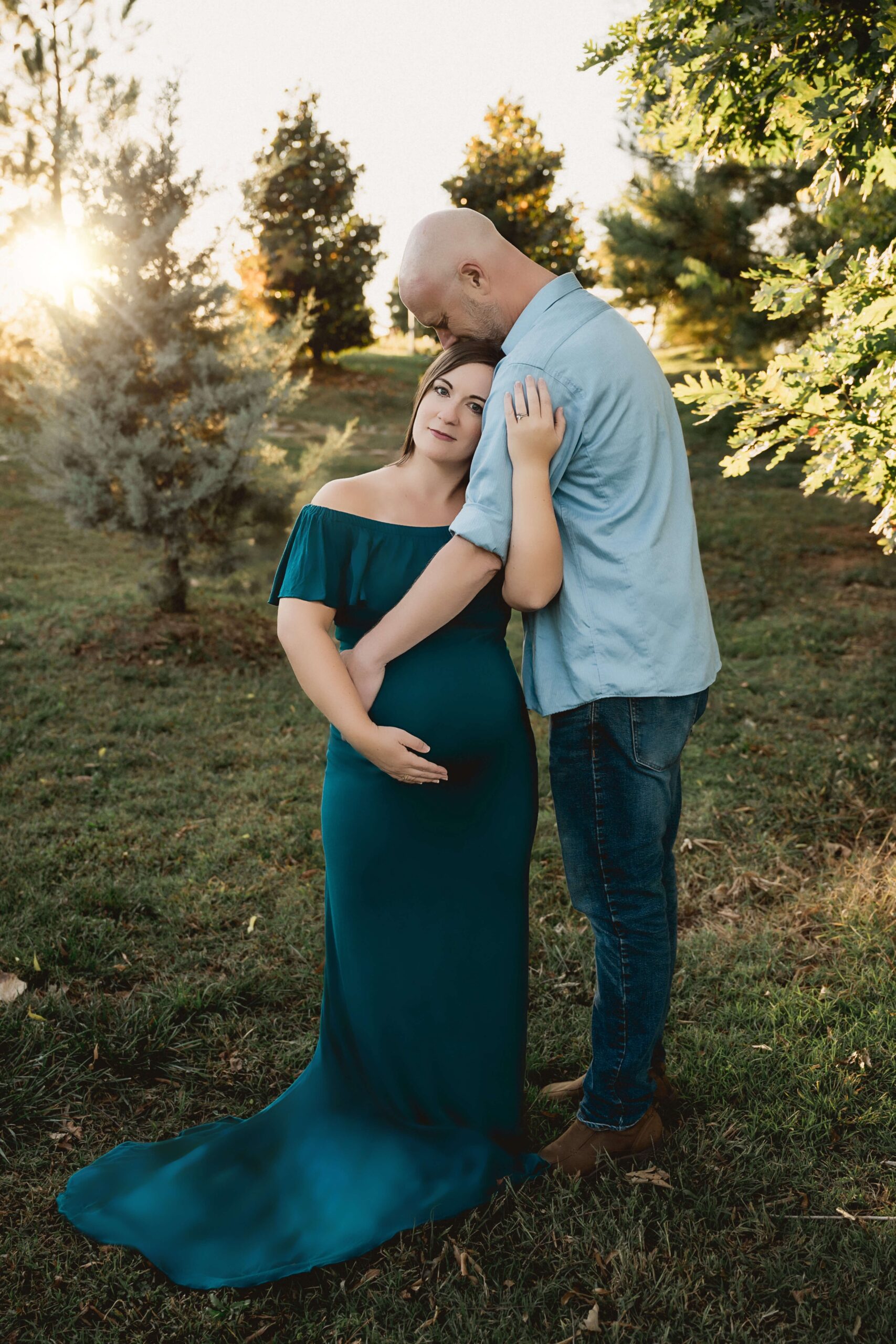 A happy expecting couple snuggles and holds the bump while standing in a park lawn at sunset during some raleigh date night ideas