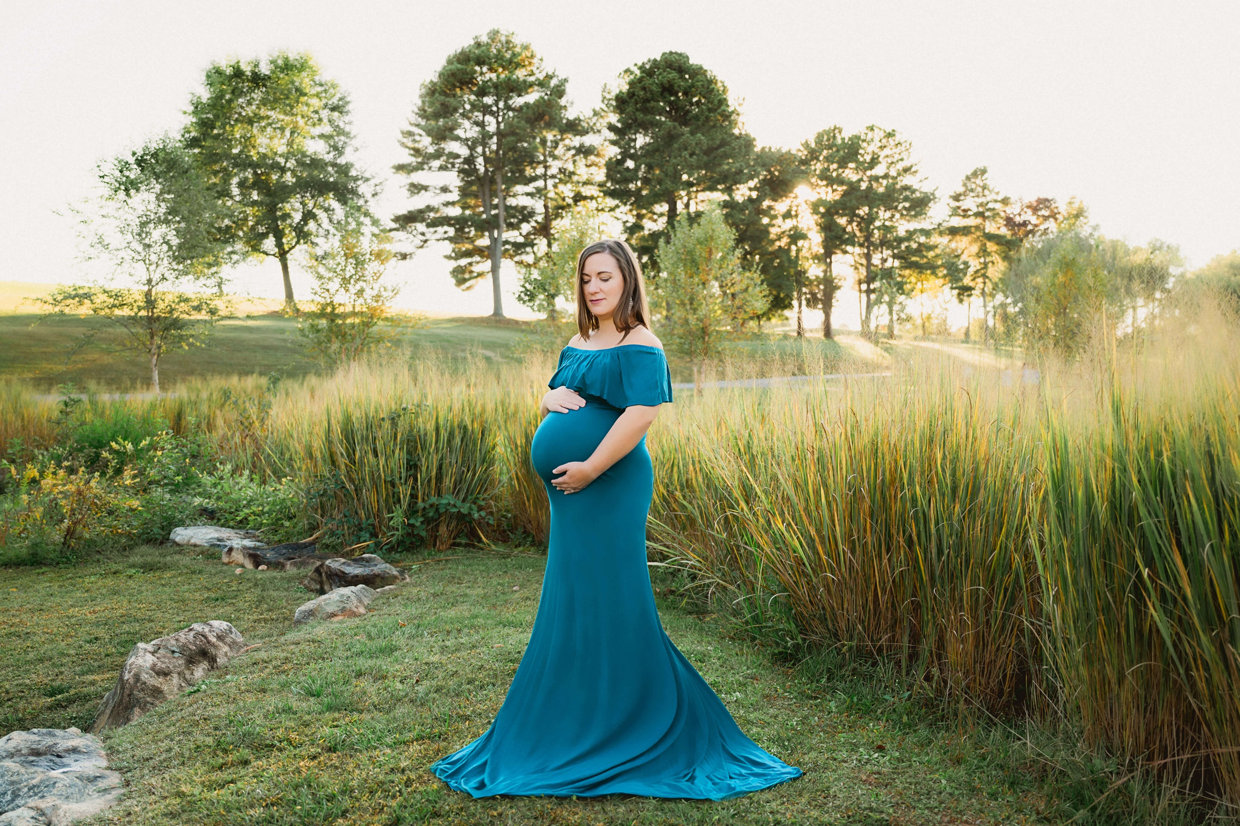 A mother to be stands in a park with tall grasses at sunset in a blue maternity gown with hands on her bump before some raleigh date night ideas