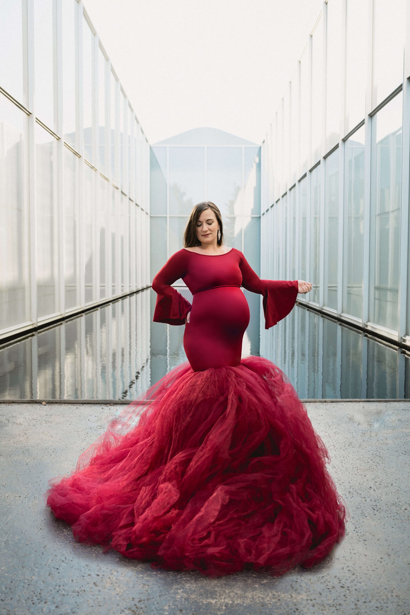 A mother to be standing by an indoor pond with an arm out in a flowing red maternity gown