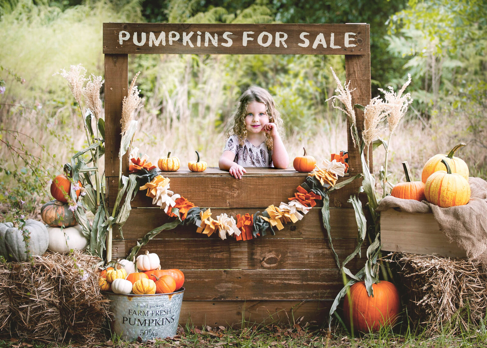 A toddler girl stands in a wooden pumpkin stand surrounded with pumpkins and corn during raleigh fall activities