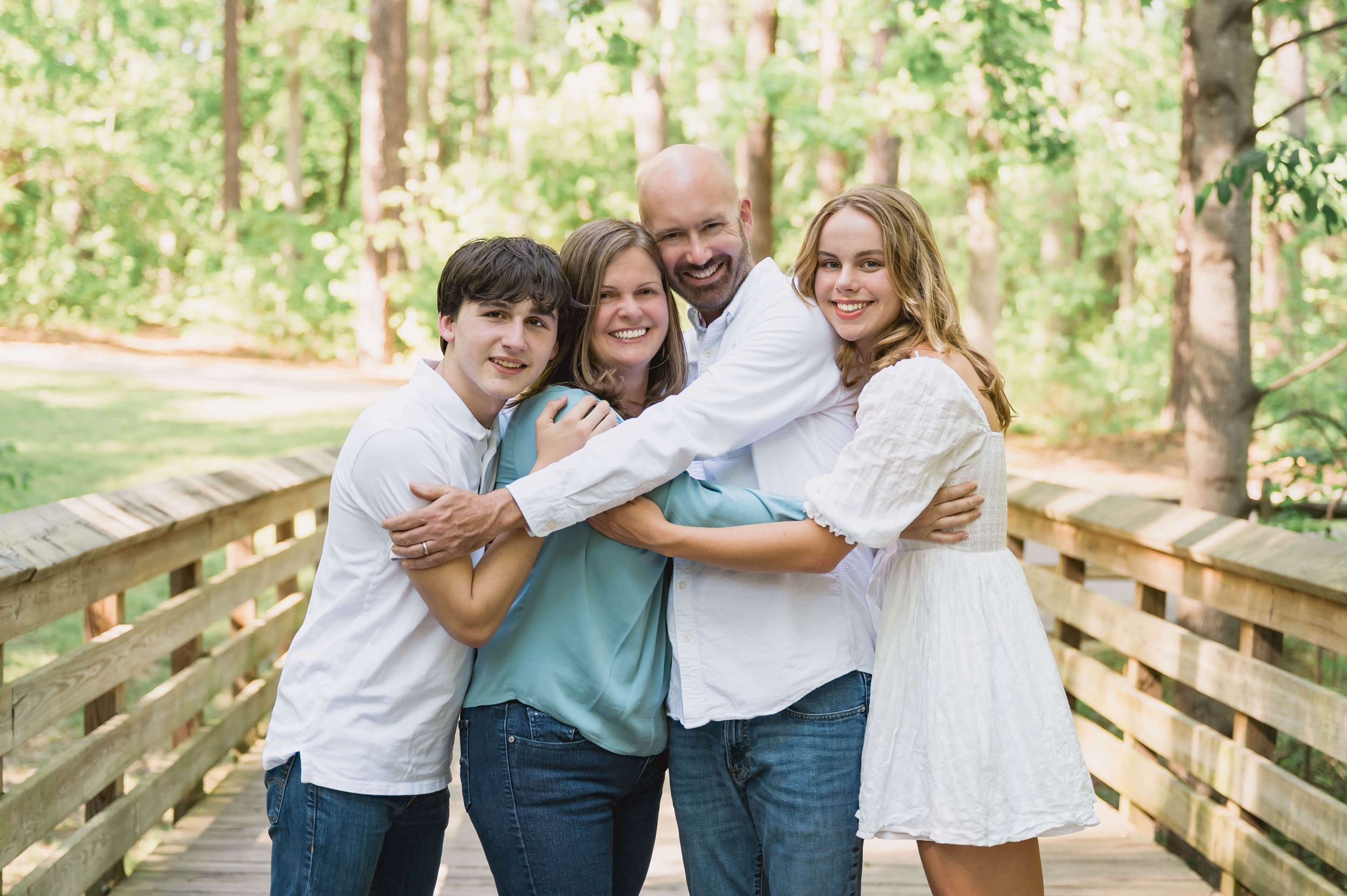 A happy family of 4 hug and smile while standing on a park boardwalk