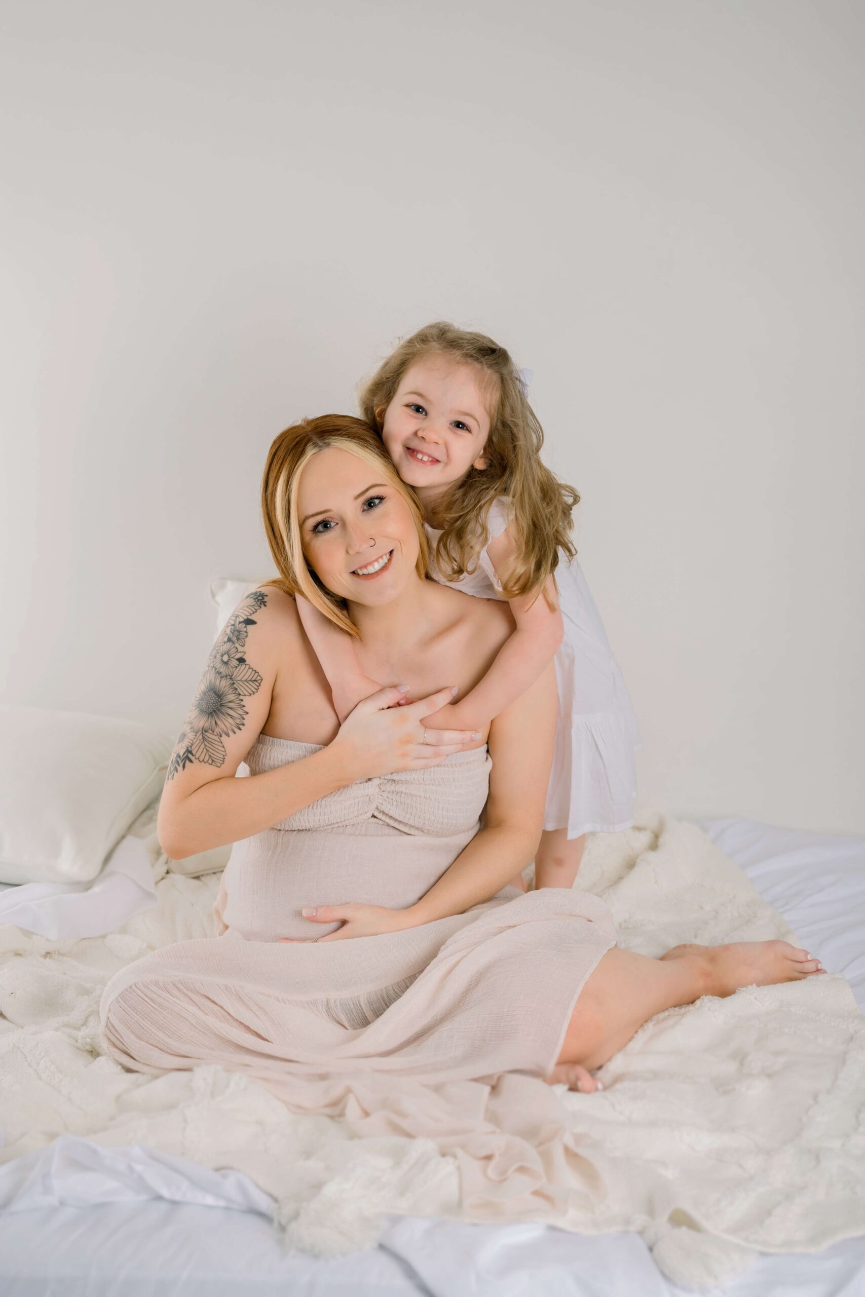 A toddler girl in a white dress hugs her pregnant mom from behind while they sit on a bed before visiting a raleigh obgyn