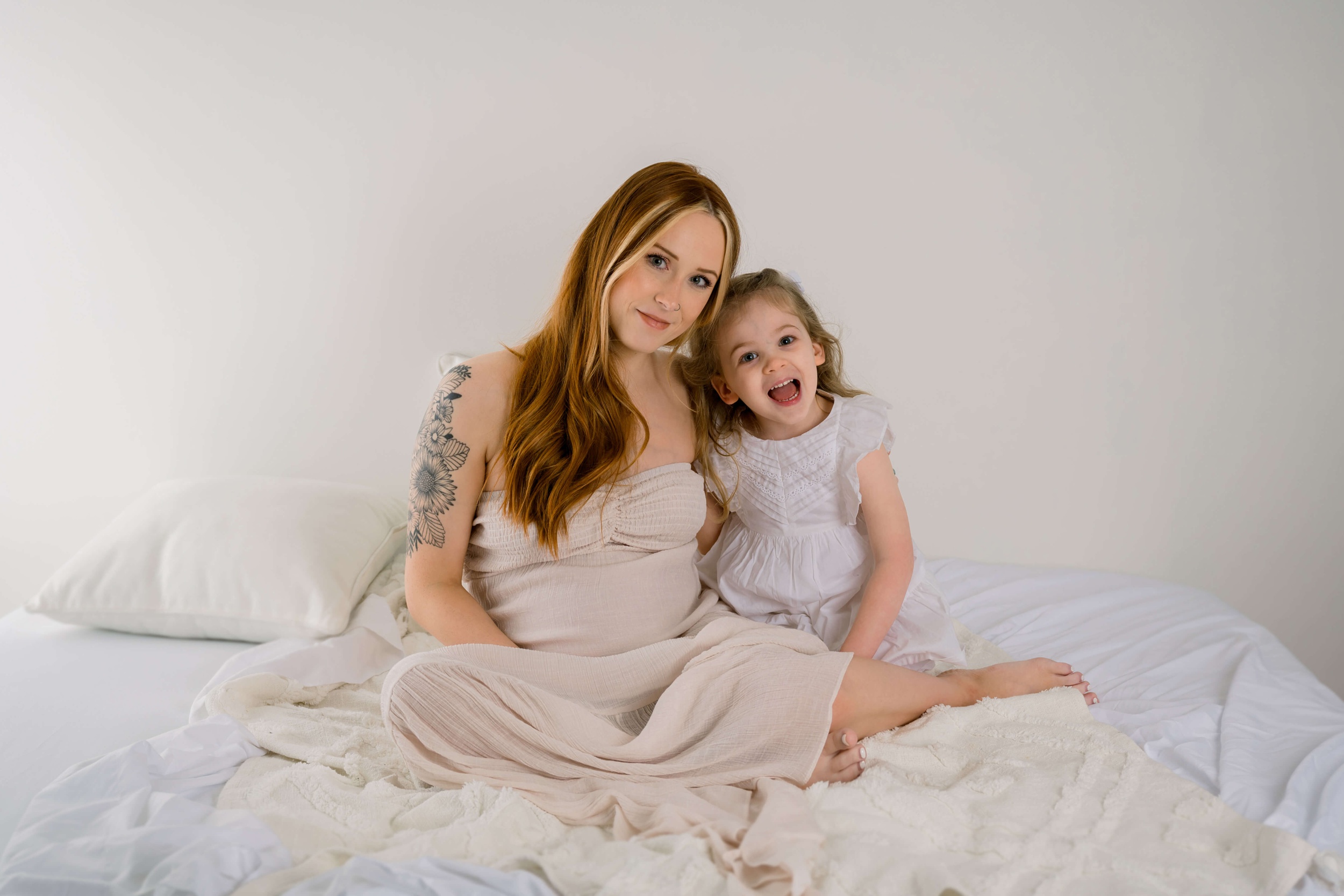 A toddler girl laughs while sitting with mom on a bed in a studio before mom visits a raleigh obgyn