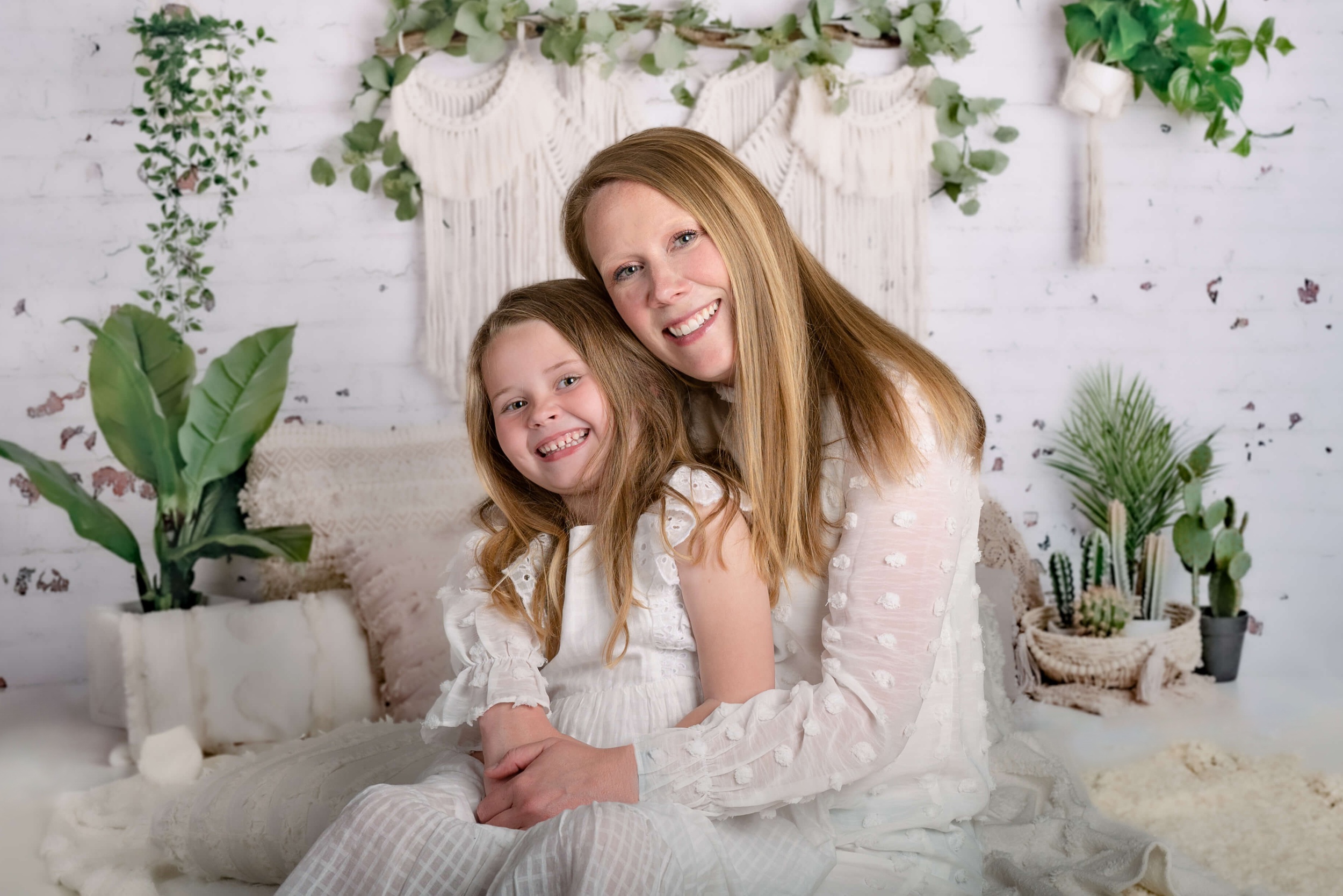 A happy mom and daughter sit on a bed together smiling in white dresses