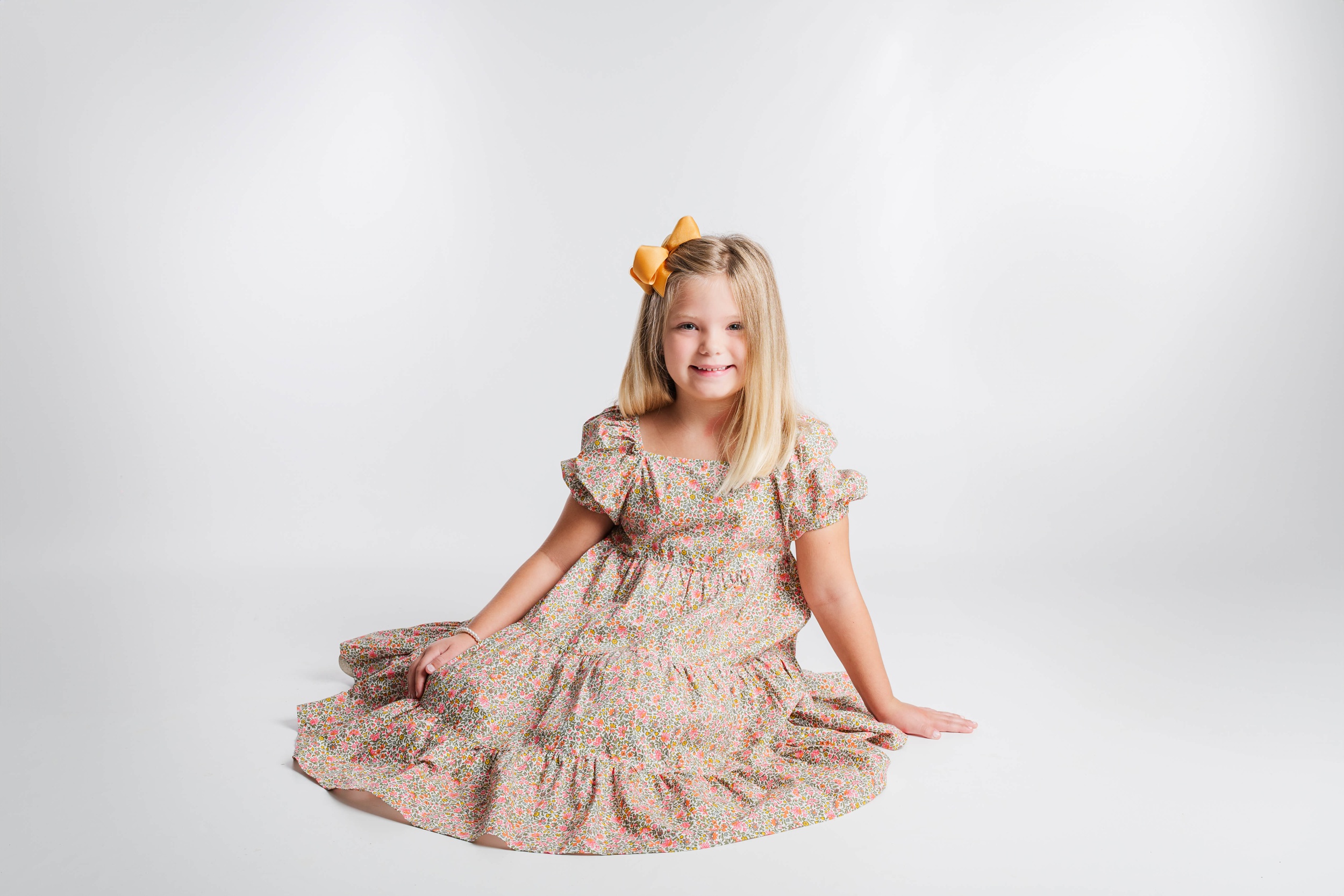 A toddler girl sits on the floor of a studio smiling in a pink floral print dress