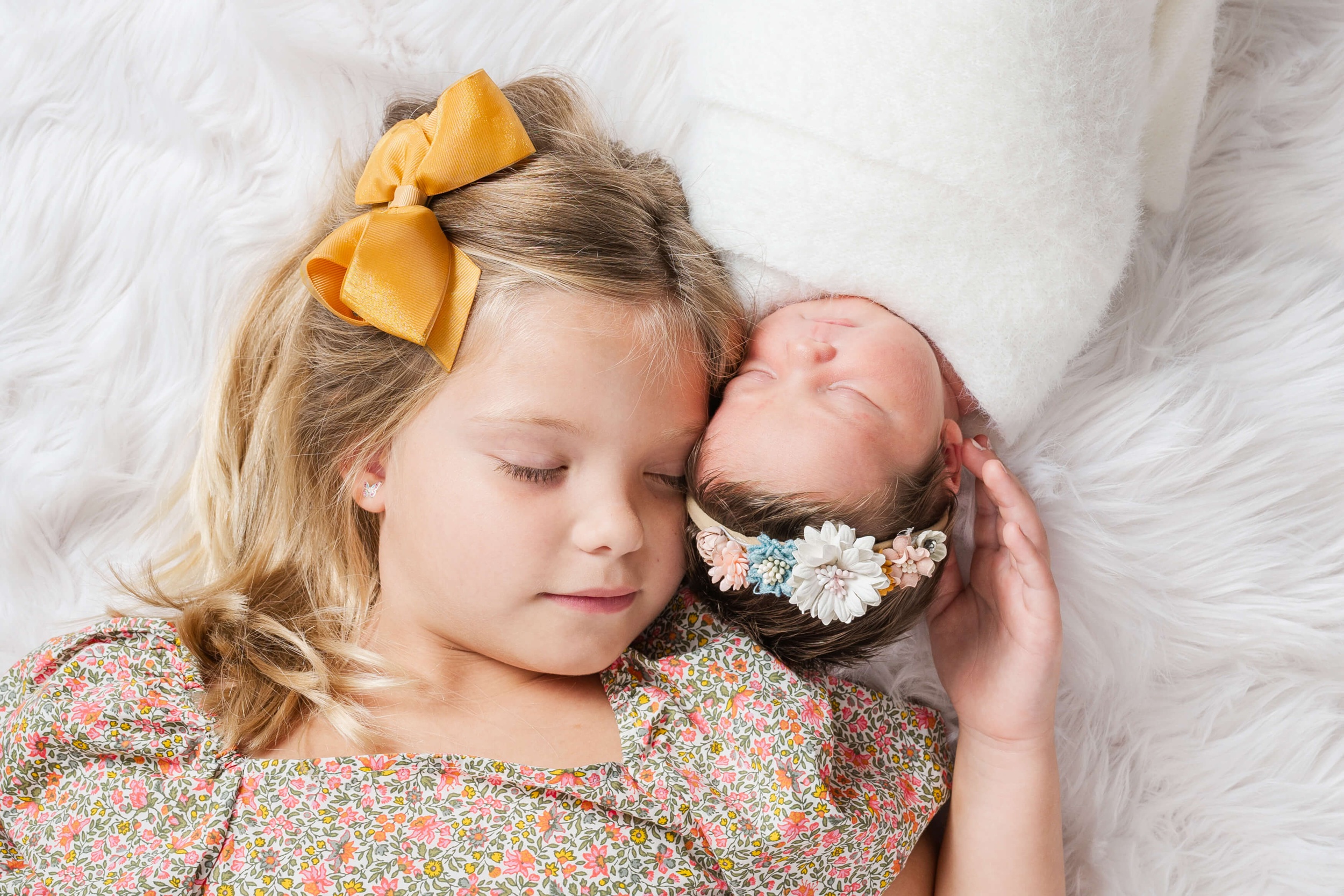 A toddler girl with a yellow bow snuggles her sleeping newborn baby sister on a shag blanket