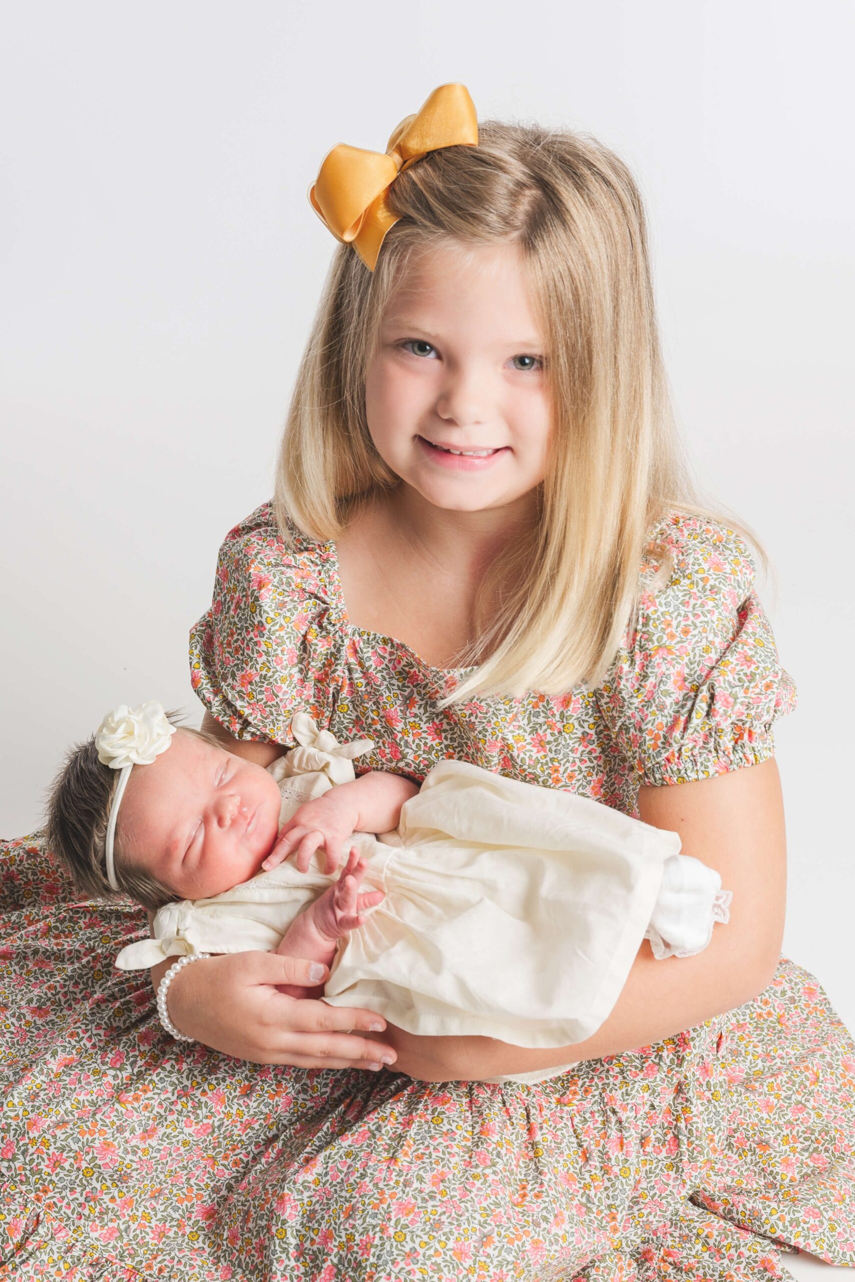 A young toddler girl in a pink floral print dress holds her newborn baby sister in her lap during things to do in raleigh nc