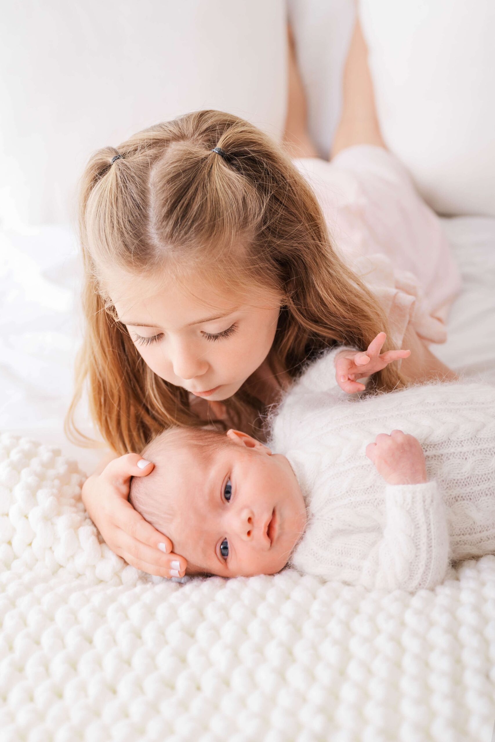 A newborn baby lays on a white bed in a white onesie while big sister lays with it