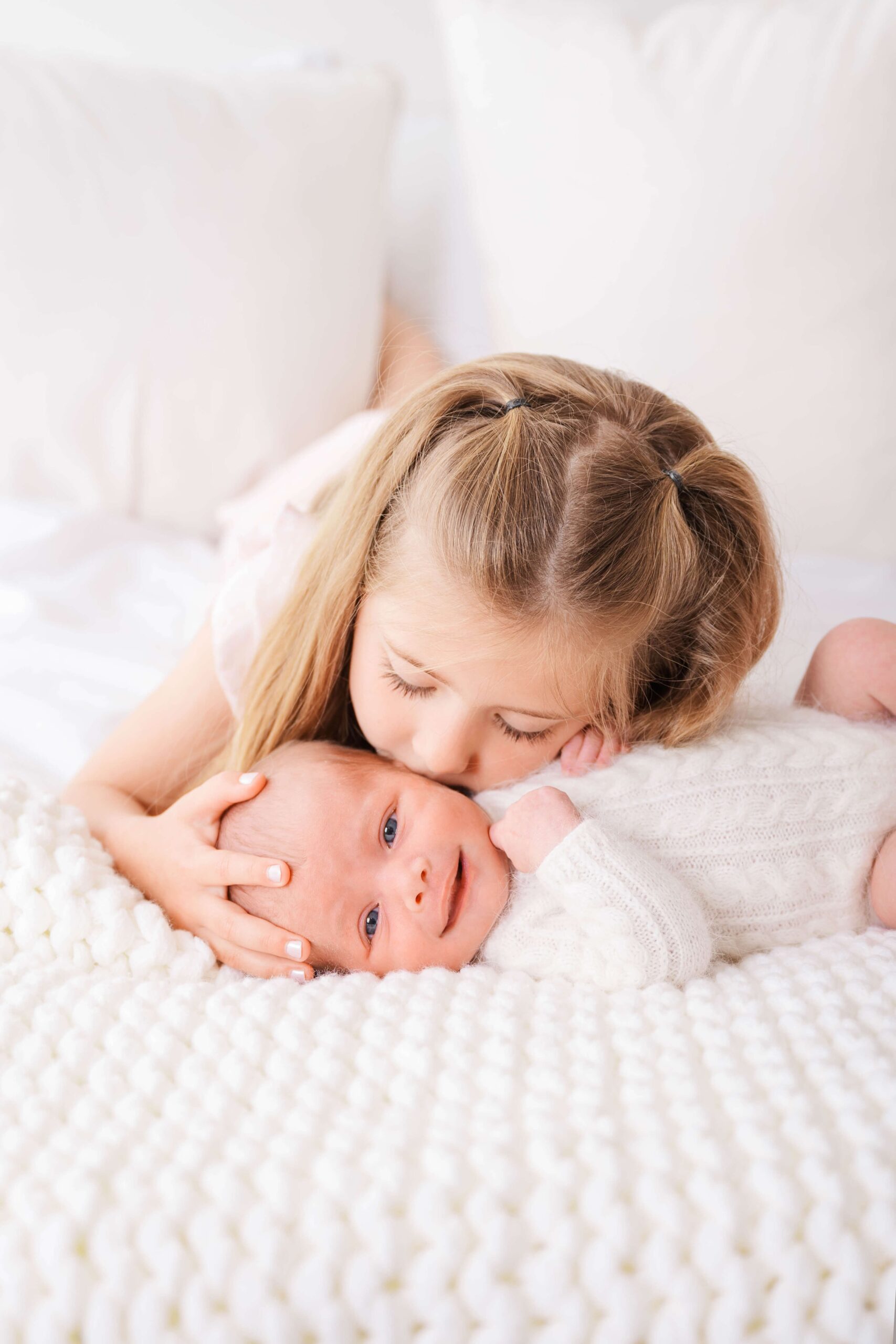 A toddler girl kisses the cheek of her smiling newborn sibling on a white bed during things to do in raleigh nc