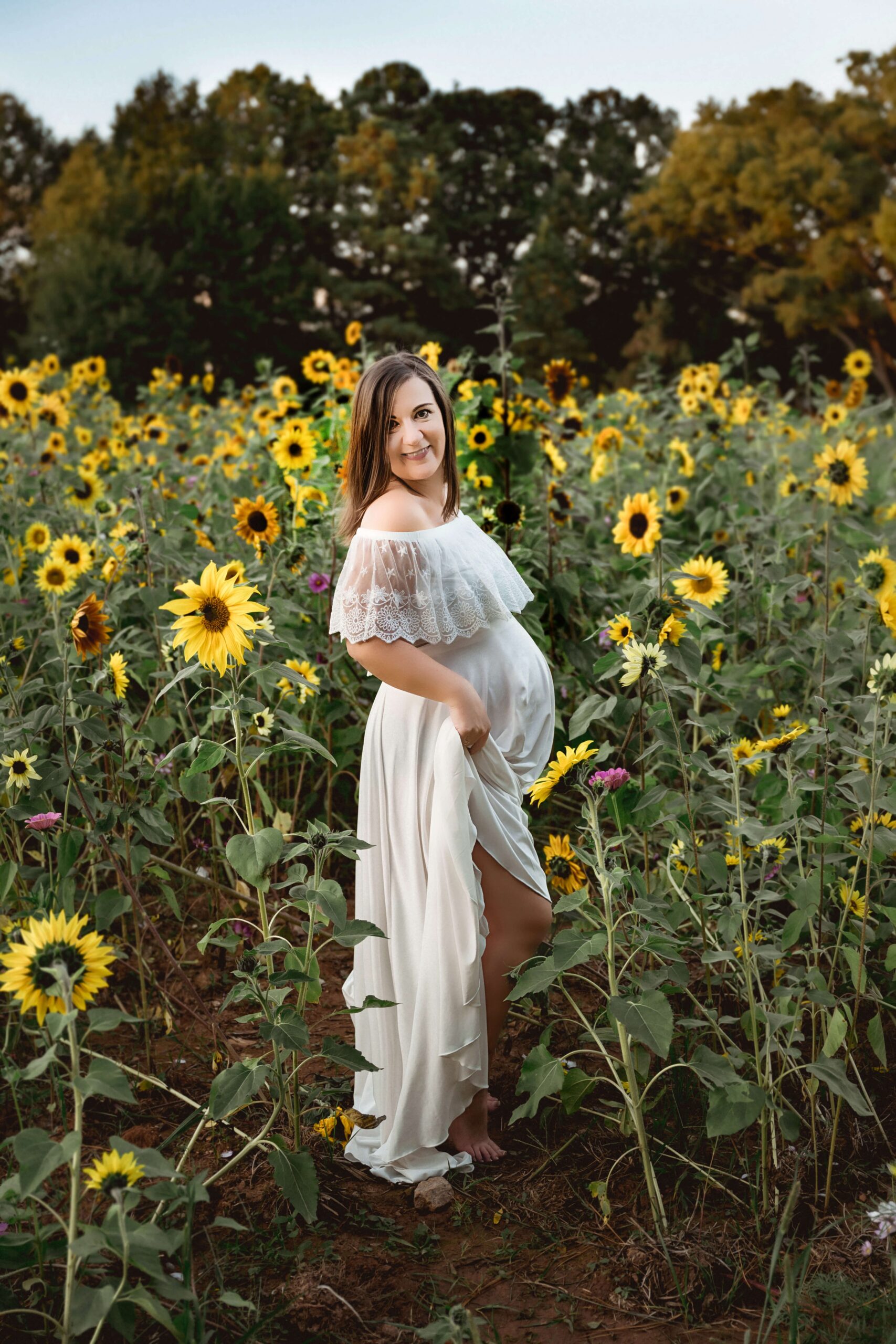 A pregnant woman smiles over her shoulder while holding her white dress train in a field of sunflowers at sunset after visiting unc rex women's center