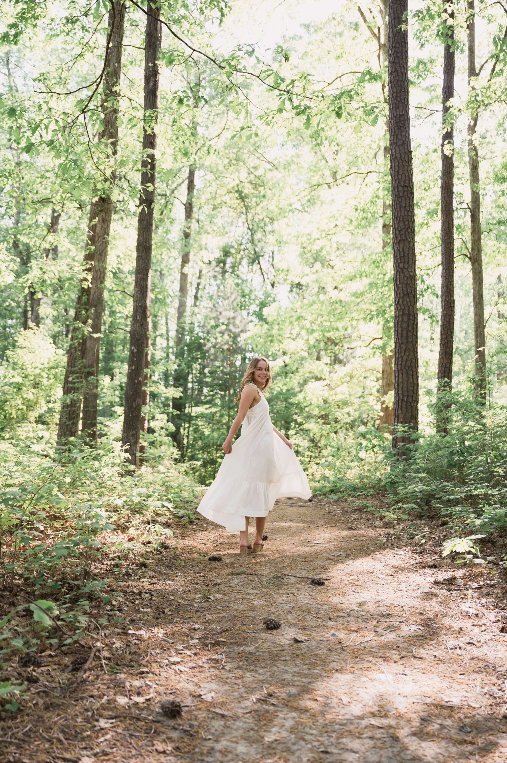 A high school senior turns while hiking in a pine forest in a white dress